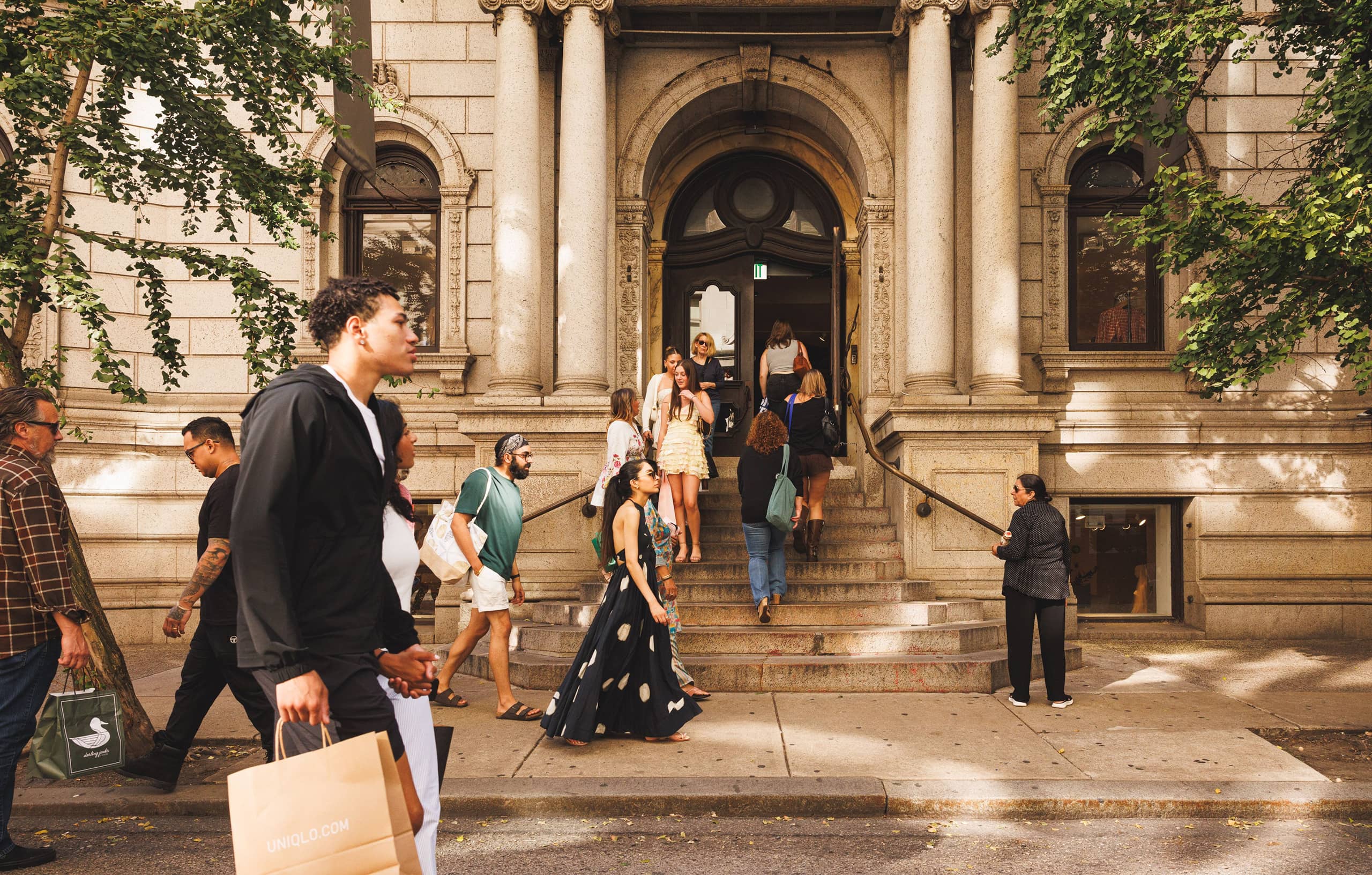Pedestrians walk past Anthropologie's grand building entrance. People are ascending the steps and others are walking by with shopping bags.
