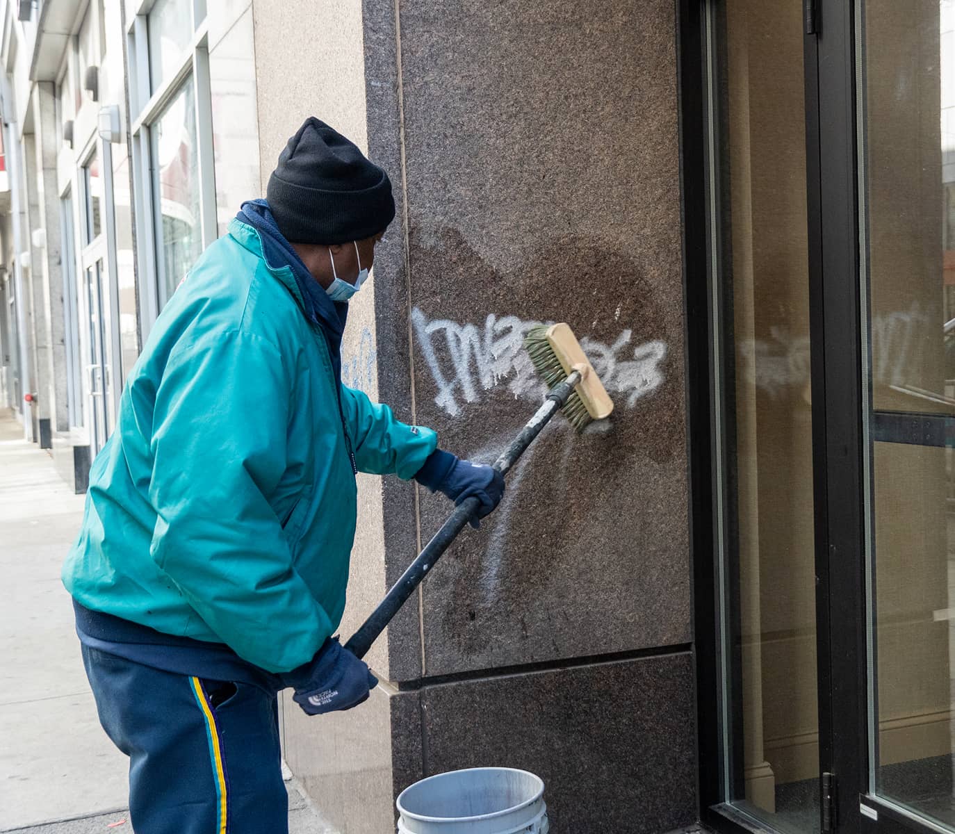 A CCD maintenance staff person in a teal jacket and black hat is scrubbing graffiti off a brown stone wall with a brush.