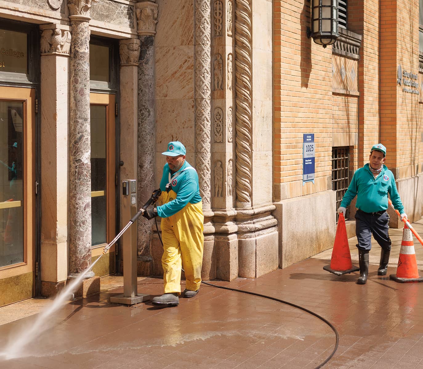Two CCD workers in teal uniforms and caps clean a sidewalk outside an ornate brick building. One uses a power washer, while the other sets up orange cones.