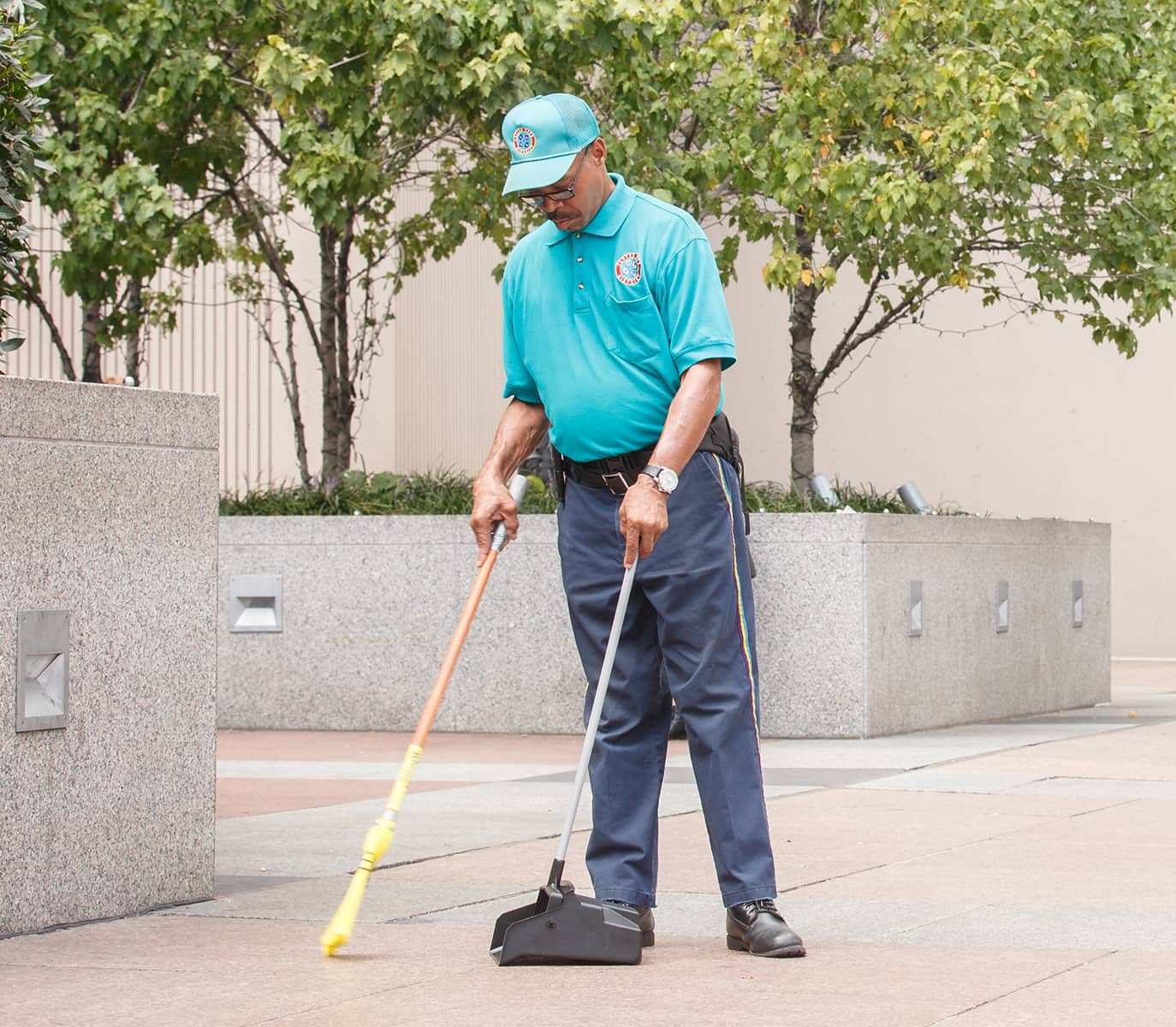 CCD maintenance staff person sweeps a plaza lined with trees and stone planters. He wears a teal uniform and cap, focused on his task.