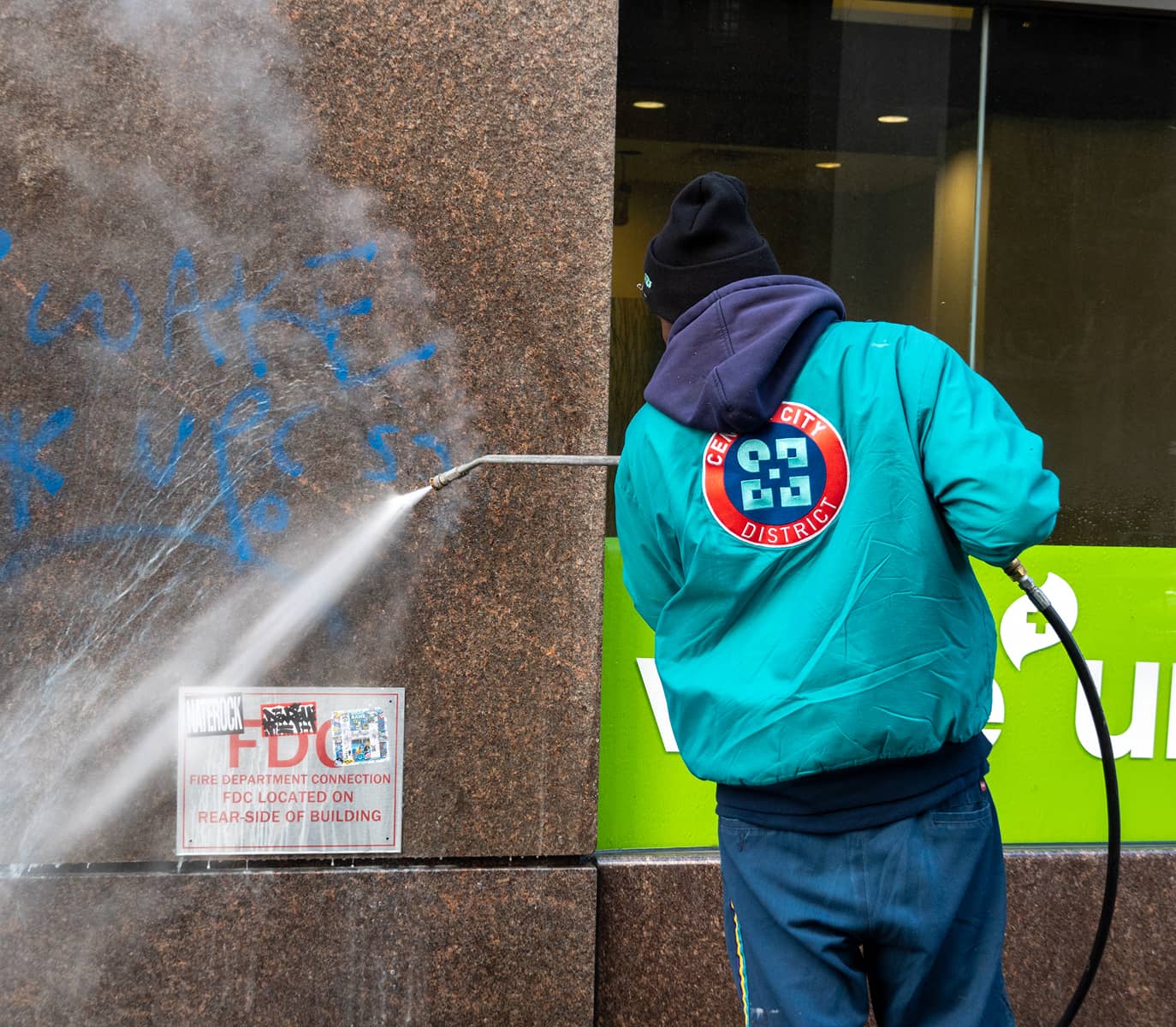 A CCD maintenance staff person in a blue-green jacket power washes blue graffiti off a brown stone wall. The jacket has a red circular logo. The scene conveys urban cleanup.