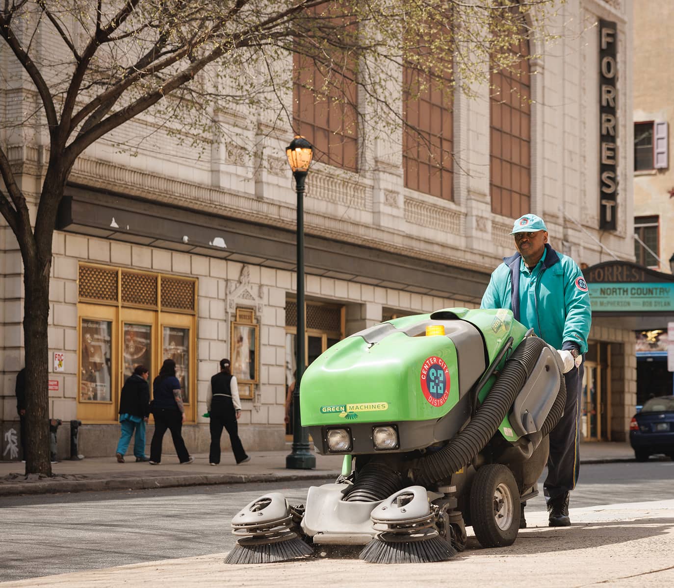 A CCD street cleaner operates a green machine, sweeping a city sidewalk. Nearby buildings and a theater marquee are visible, with pedestrians walking by.
