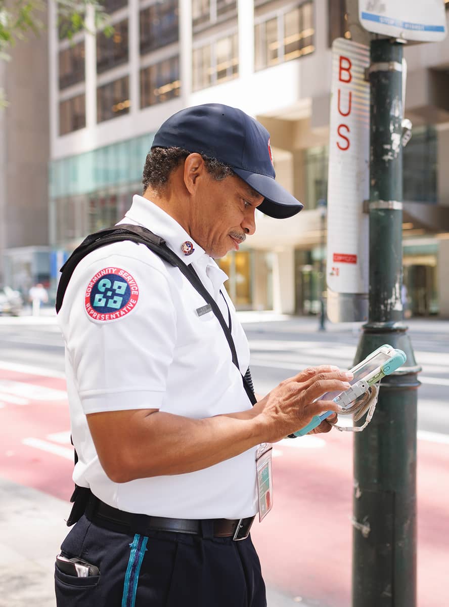 A CCD community service representative (CSR for short) safety ambassador wearing a uniform and carrying a 2-way radio, reporting a streetscape issue on a tablet.