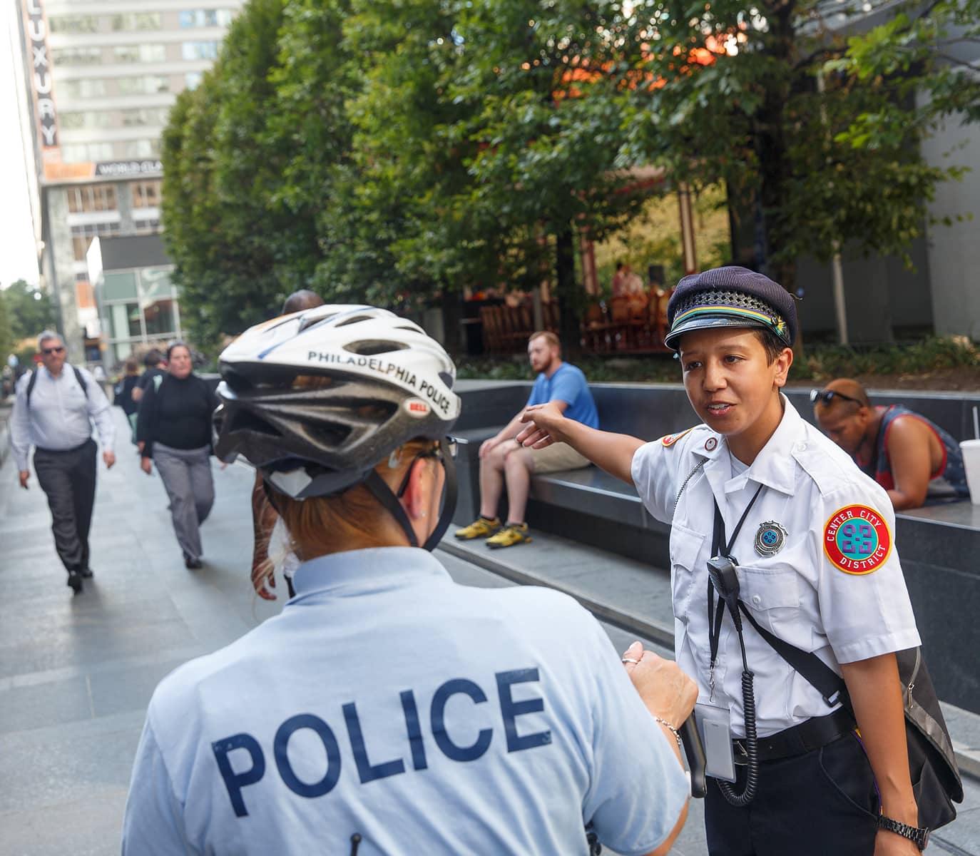 Police officers engage in a conversation on a city sidewalk with a CCD community service representative (CSR for short) safety ambassador. People walk by and sit on benches amid urban greenery.
