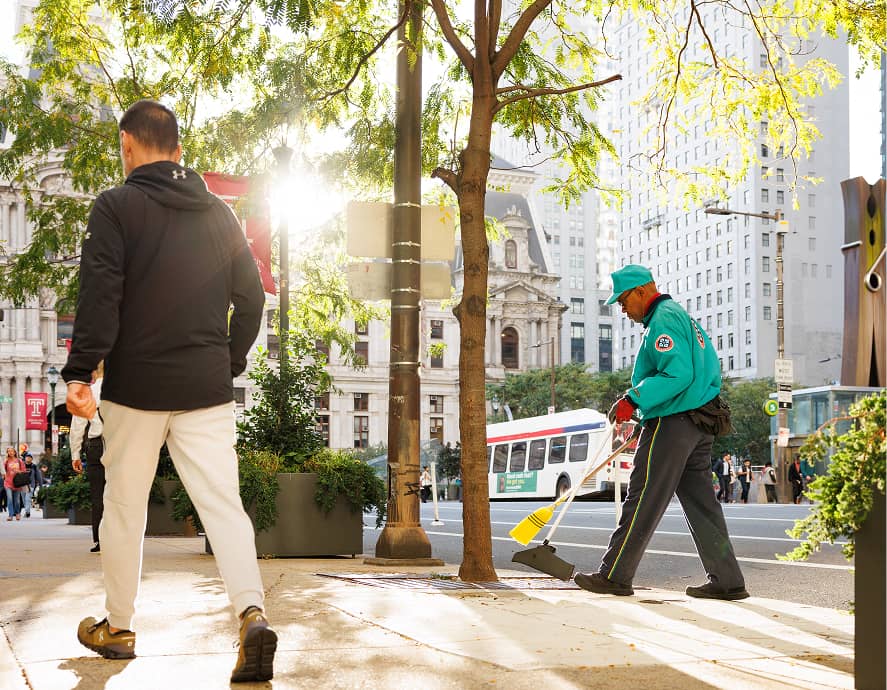 A CCD maintenance staff person in teal sweeps the sidewalk near a tree in bright daylight. A person walks by, while urban buildings and a bus are visible in the background.