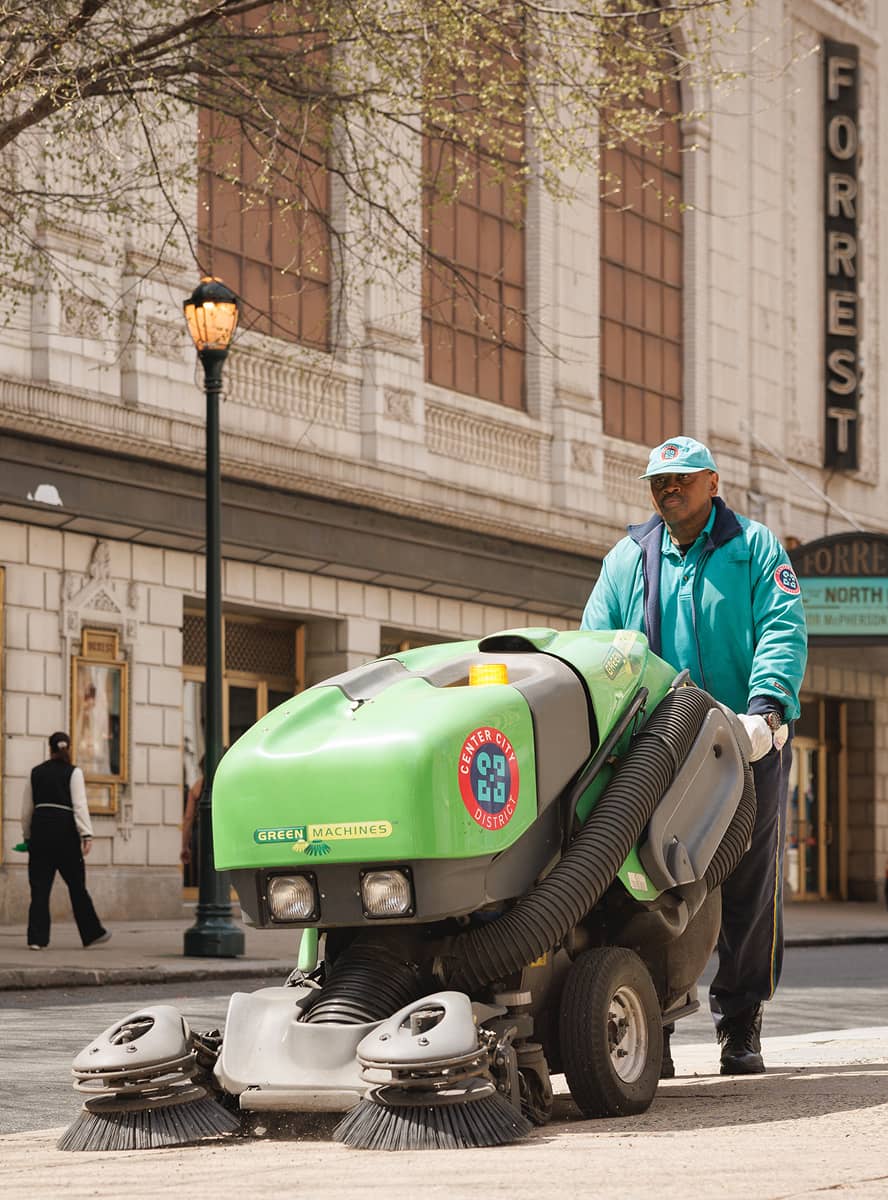 A CCD maintenance staff person operates a green machine, sweeping a city sidewalk. Nearby buildings and a theater marquee are visible, with pedestrians walking by.