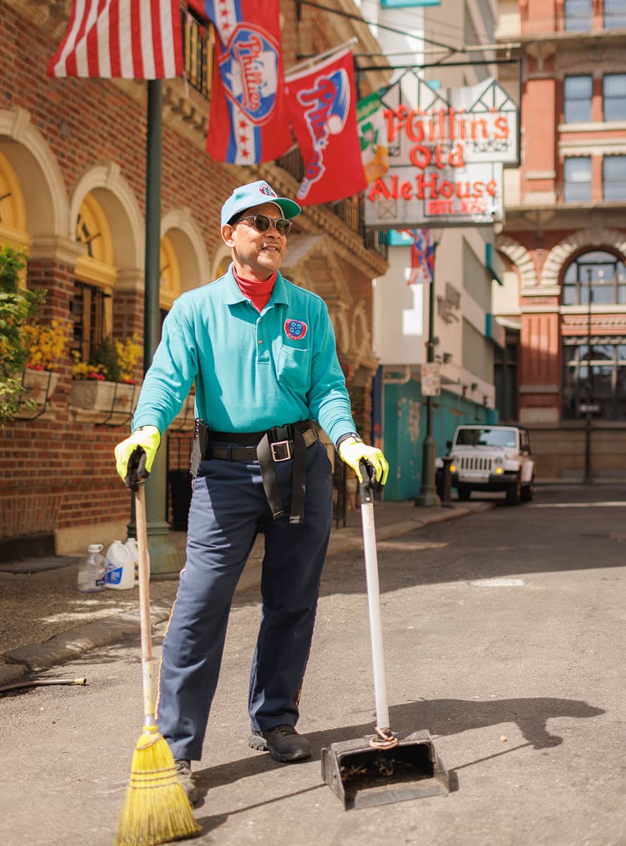 A CCD maintenance staff person in a teal uniform is smiling and sweeping a street. They hold a broom and dustpan, with McGillin's Old Ale House in the background.